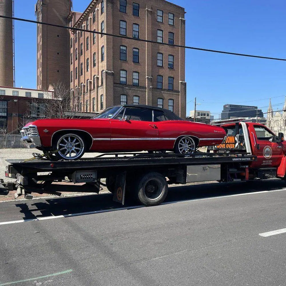 Flatbed carrying a red classic convertible in front of a brick building